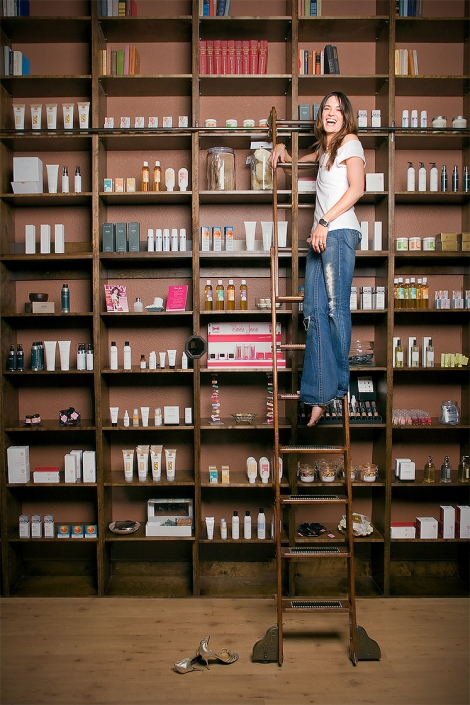 Editorial portrait of a woman in front of a wall of beauty products at the top of a book ladder with no shoes and laughing. Shot for Vegas Magazine by Phoenix editorial photographer Jason Koster.